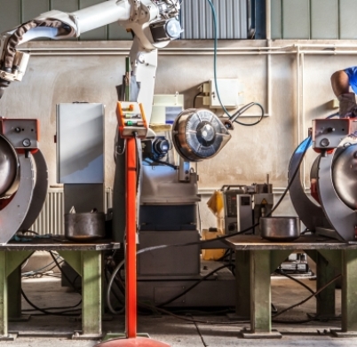 Man and robotic machine work together inside industrial building. The mechanical arm performs welds on metal components assisted by a worker who in turn manages welds manually.